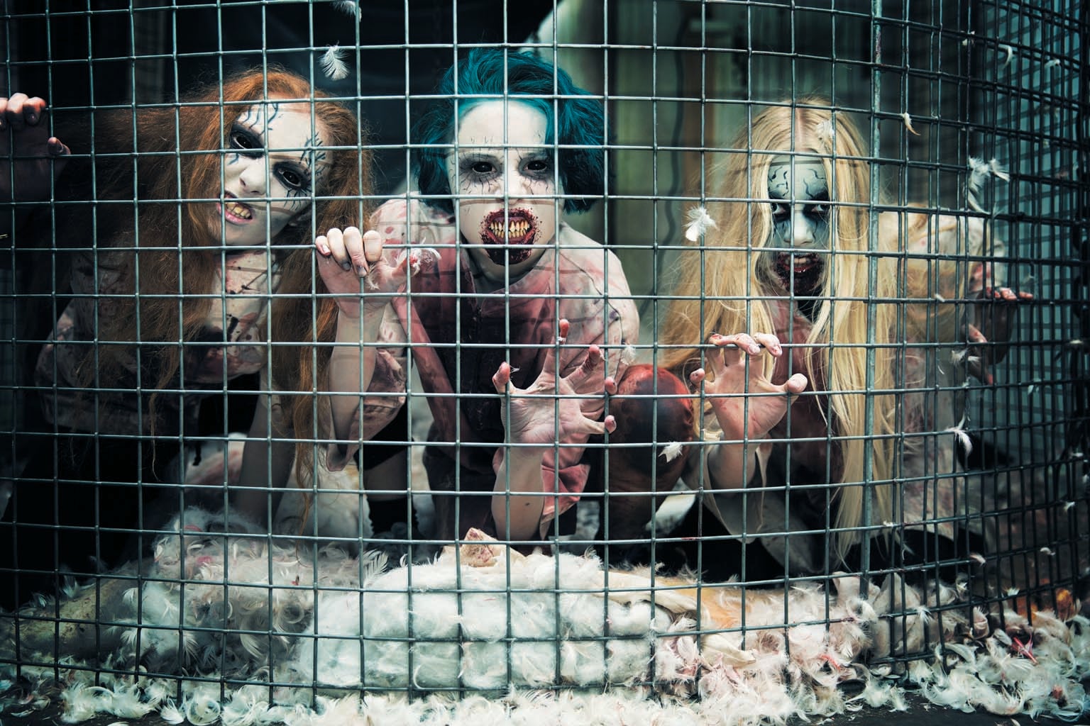 Three people dressed as horror creature with white painted faces and darkened eyes, shown behind a wire fence.